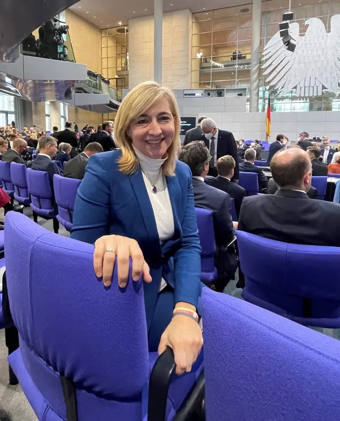 Nina Warken smiling in the plenary chamber of the German Bundestag, rows of blue seats behind her