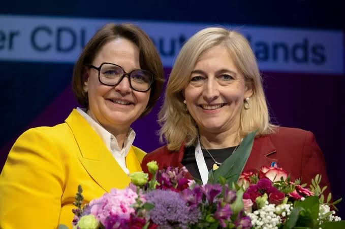 Nina Warken smiling beside a woman in a yellow jacket at a CDU event, with flowers in the foreground