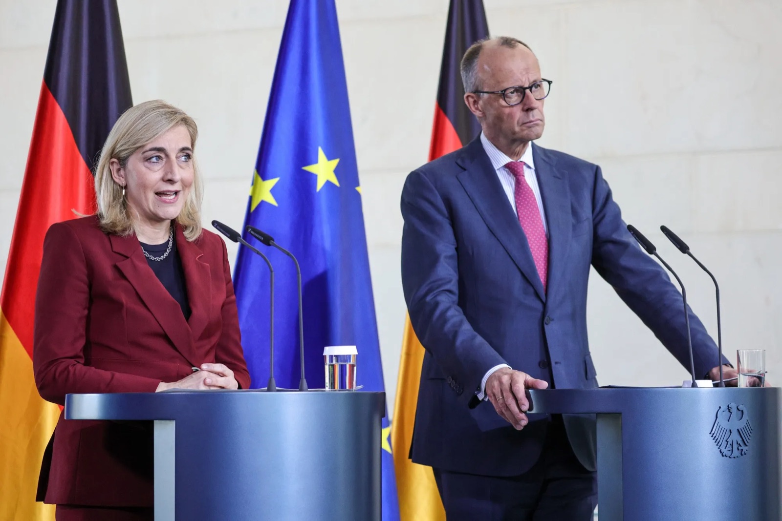 Nina Warken in a dark red suit speaking at a lectern beside Chancellor Friedrich Merz, with German and European flags behind them