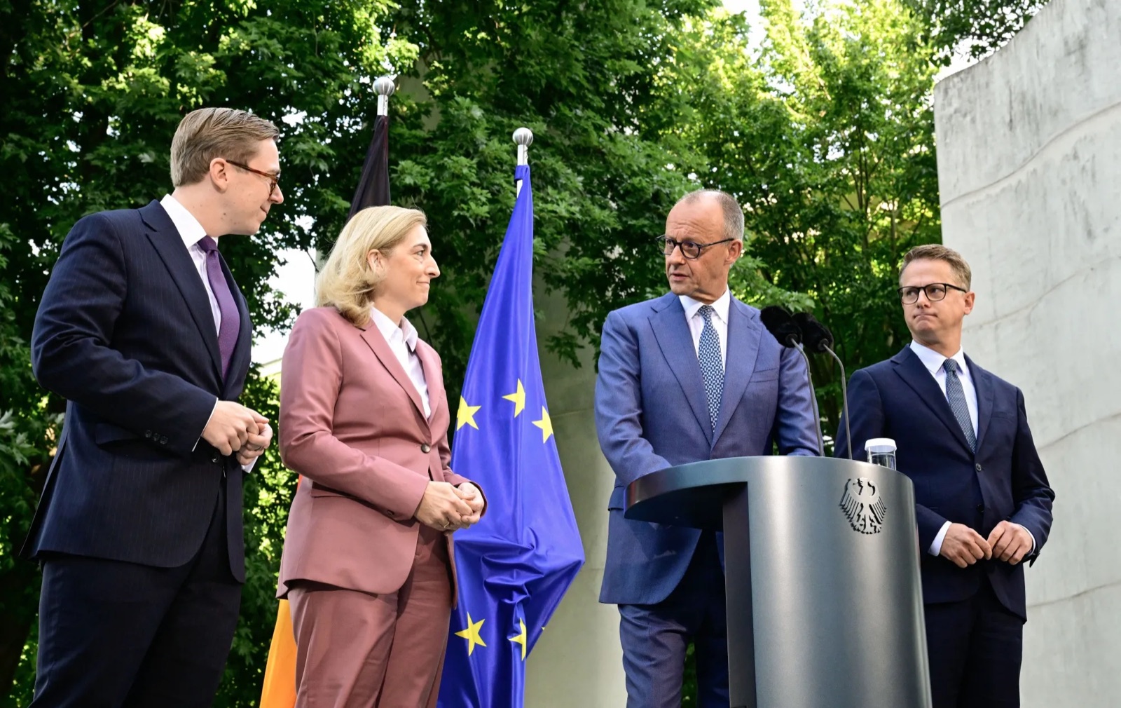 Chancellor Friedrich Merz speaking at an outdoor lectern; Nina Warken in a rose-coloured suit and two men stand beside him in front of trees and German and European flags
