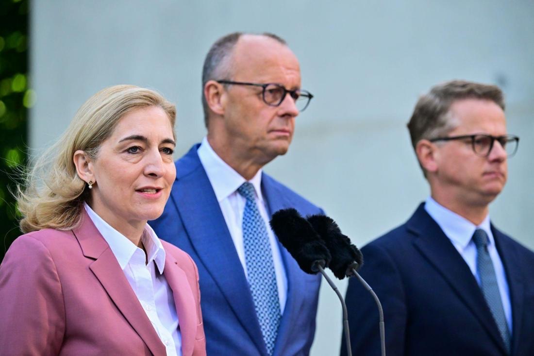 Nina Warken in a pink blazer speaking into microphones, with Chancellor Friedrich Merz and Carsten Linnemann standing behind her