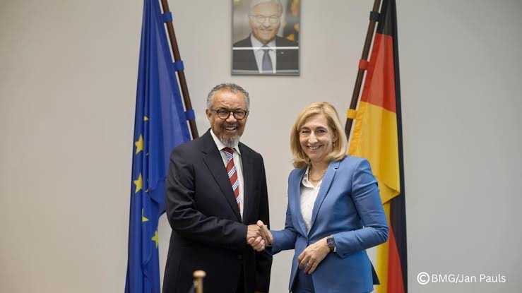 Nina Warken in a light blue suit shaking hands with WHO Director-General Tedros Adhanom Ghebreyesus in front of European and German flags