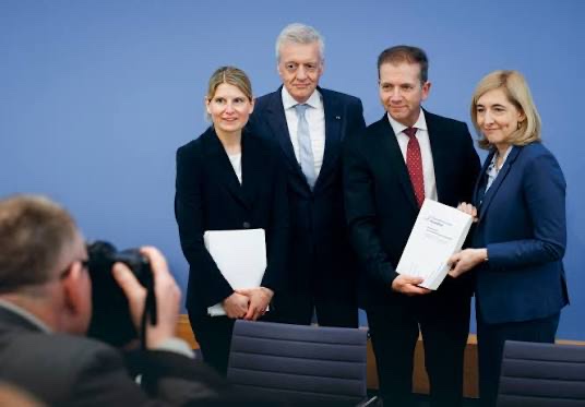 Nina Warken holding up a printed report at a press conference, standing with three other people in front of a blue backdrop