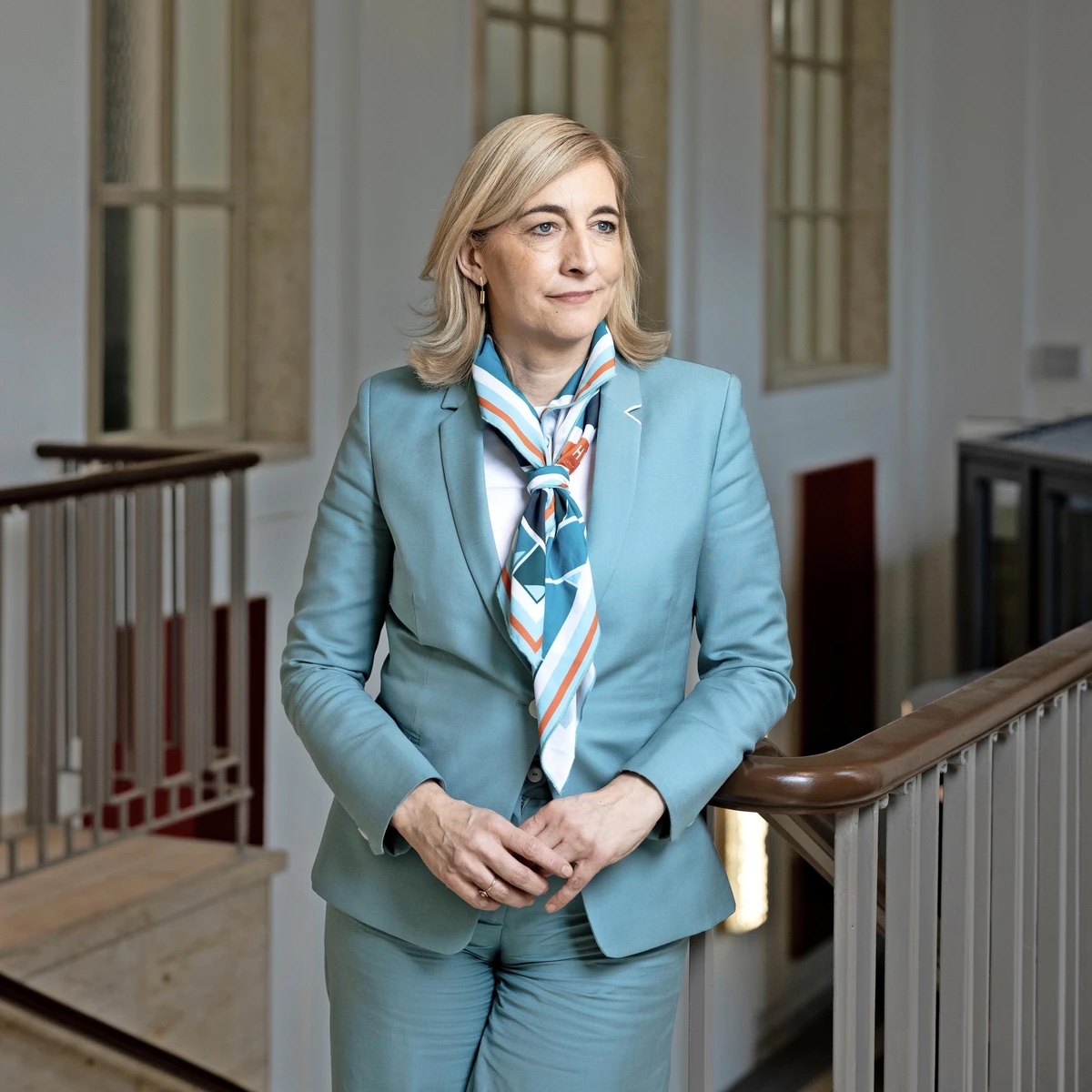 Nina Warken, full-length portrait in a light blue suit and patterned scarf, standing beside a stair banister