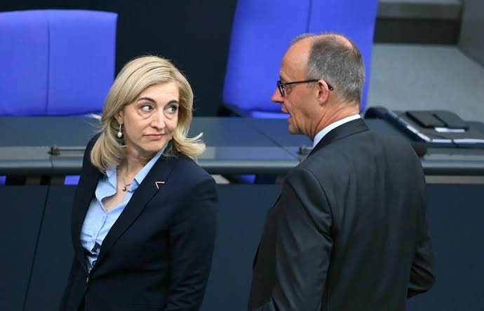 Nina Warken and Chancellor Friedrich Merz standing in conversation in front of the blue seats of the Bundestag plenary chamber
