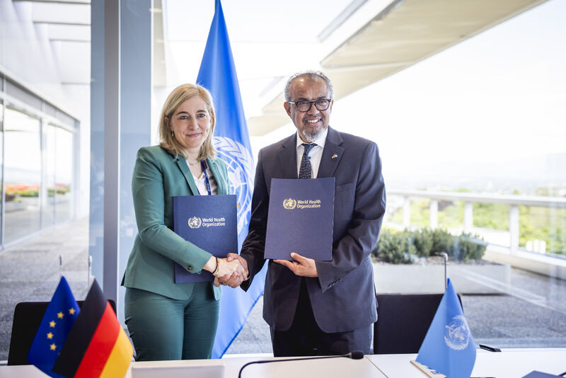 Nina Warken in a green suit shaking hands with WHO Director-General Tedros Adhanom Ghebreyesus; both hold blue World Health Organization folders, with WHO, EU and German flags