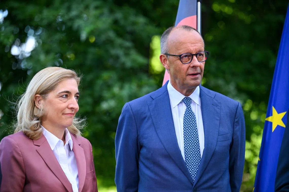 Nina Warken in a rose-coloured blazer standing outdoors beside Chancellor Friedrich Merz, with German and EU flags and trees behind them